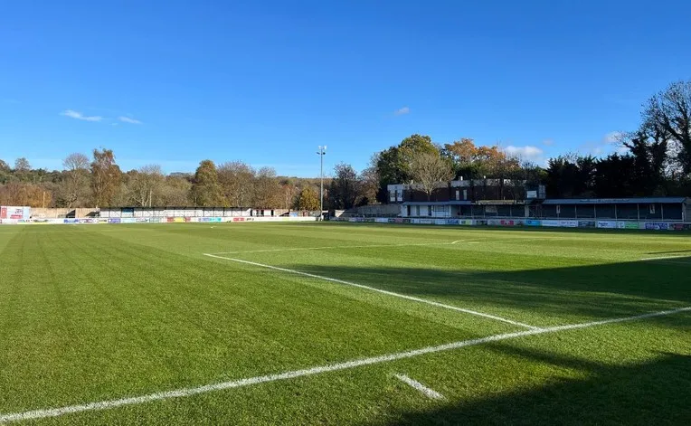 Góc nhìn từ khán đài xuống sân The Meadow Chesham United, cho thấy mặt cỏ xanh và khung cảnh sân bóng đơn sơ